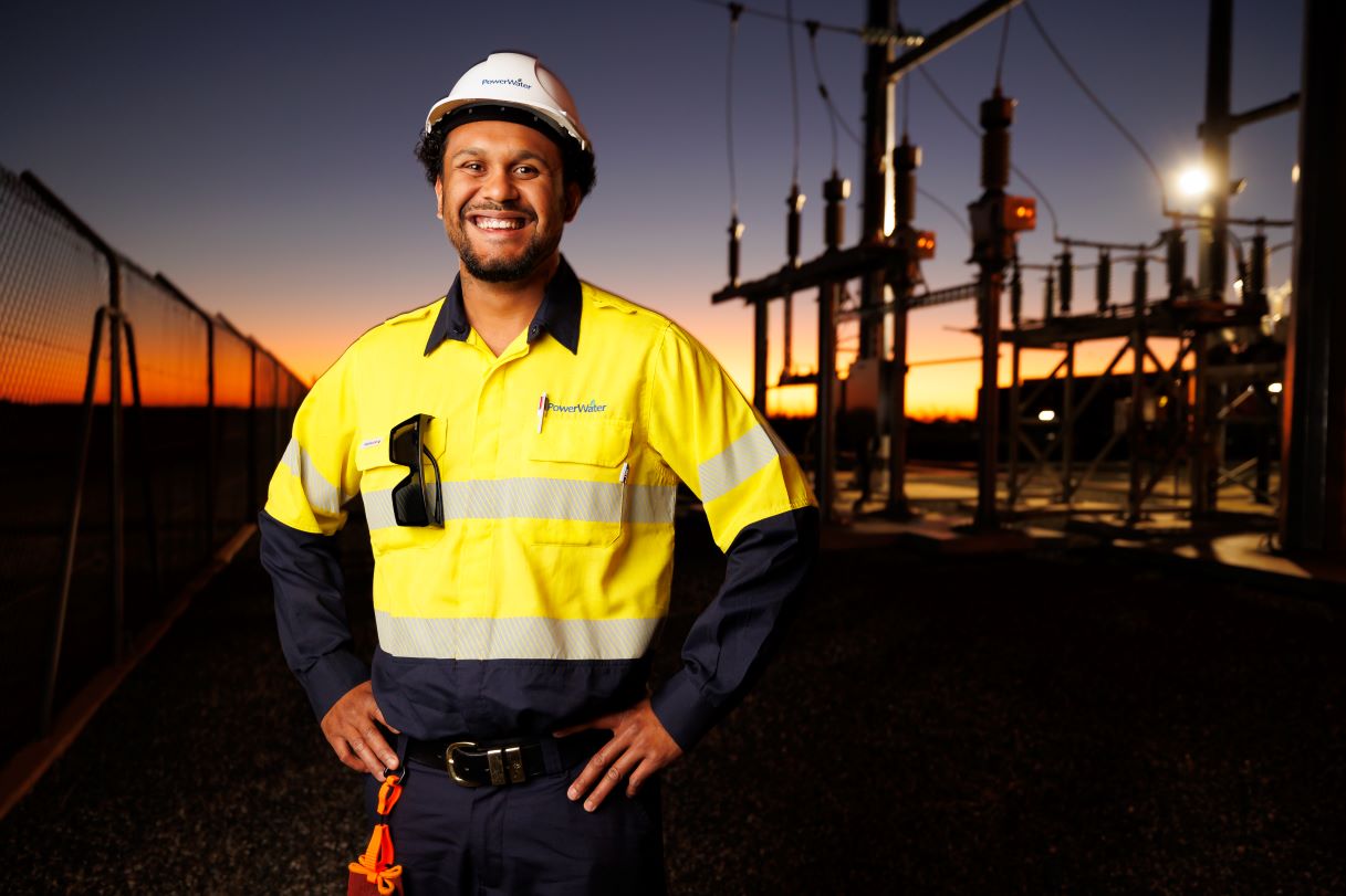 Smiling Power and Water employee wearing a yellow high-visibility uniform and a white hard hat, standing in front of an electrical substation at sunset.