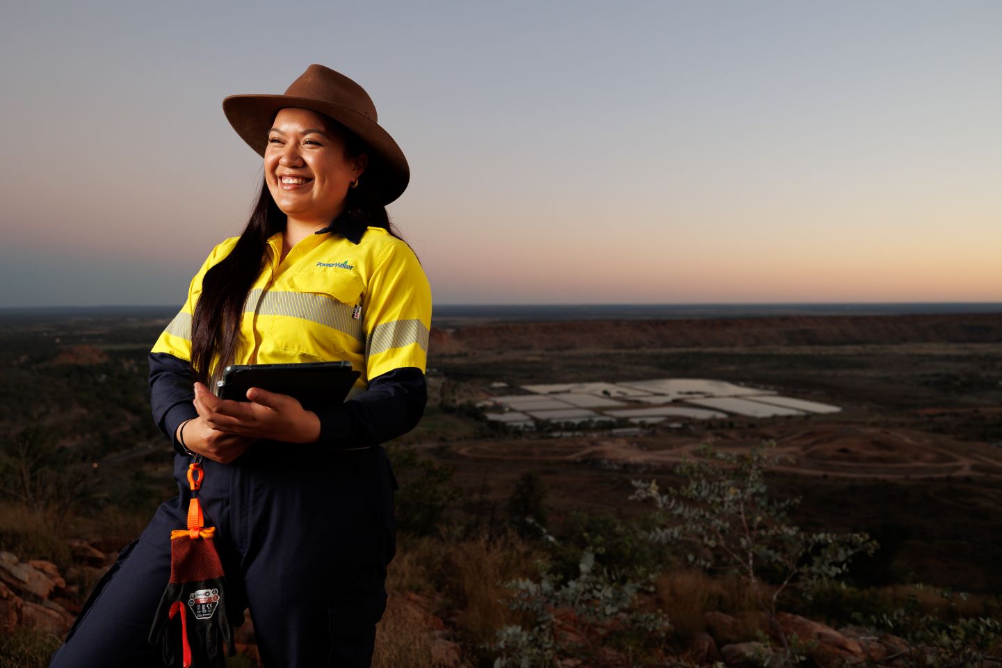 Smiling Power and Water employee wearing a yellow high-visibility in personal protective equipment uniform wearing a brown hat,  holding a tablet while standing on a hill overlooking a rural landscape at sunset.