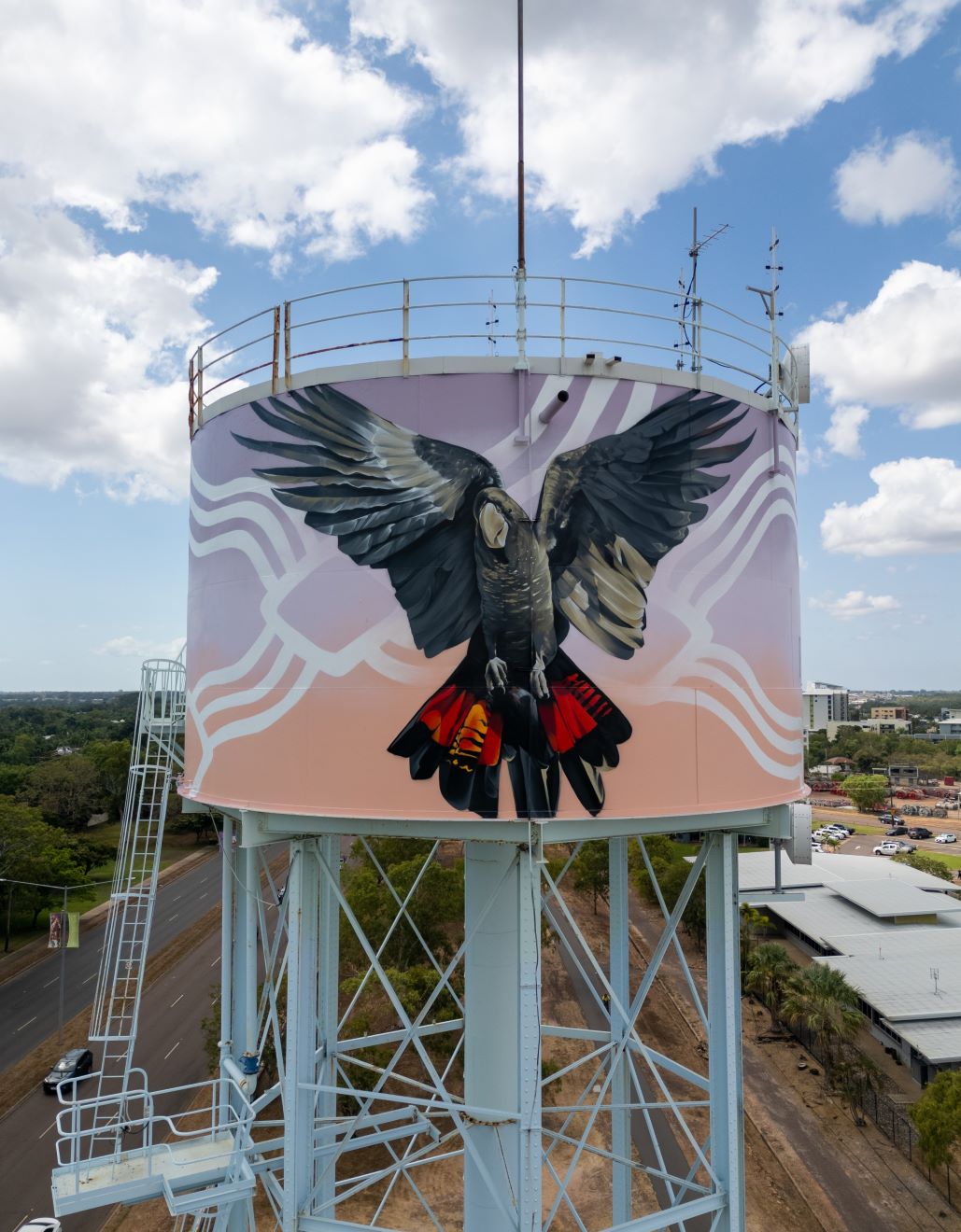  A large water tank featuring a striking mural of a black cockatoo with wings spread wide, showcasing its vibrant red and yellow tail feathers. The bird is painted in aboriginal style against a soft gradient background with pastel shades of pink, orange, and white, and abstract wavy lines.  The tank is mounted on a tall, light blue metal structure with a ladder, and the scene is set under a partly cloudy sky with nearby buildings and roads visible in the background.