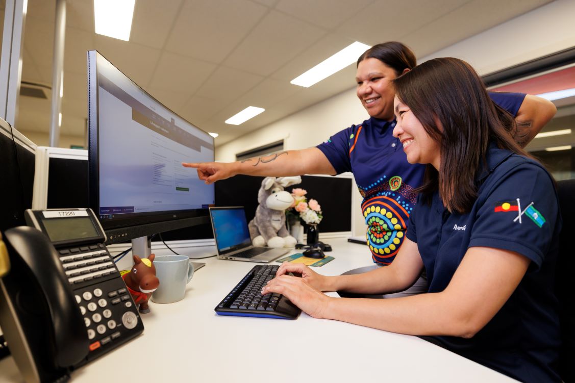 Two smiling Power and Water employees in an office setting, one sitting at a desk typing while the other stands beside them, pointing at a computer screen